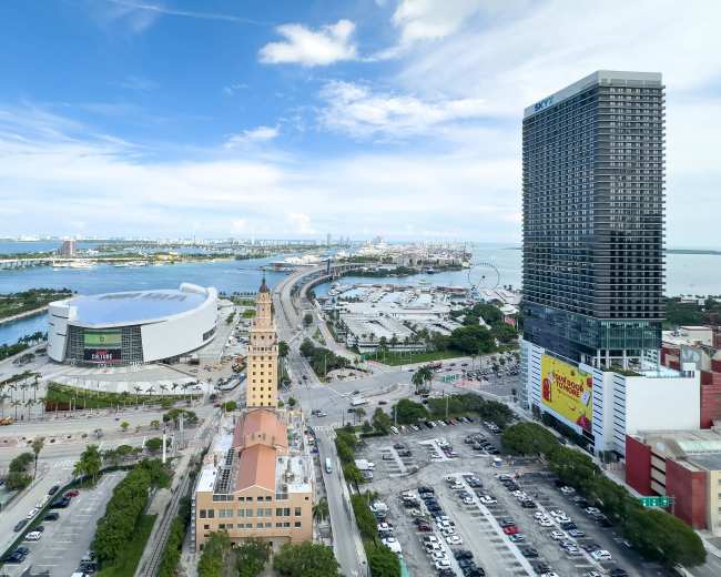 The image shows a panoramic view of a coastal city skyline, featuring streets, a sports arena, a tall building, and waterways under a partly cloudy sky.