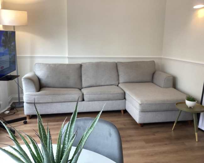 The image shows a light gray sectional sofa in a softly lit corner of a room with wooden flooring and a small table beside it.