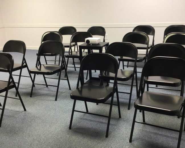 A room with several black folding chairs arranged around a central table featuring a projector.