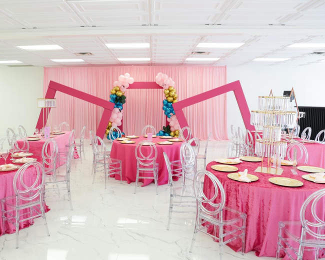 The image shows a decorated event space featuring round tables with pink tablecloths and clear acrylic chairs, arranged under a colorful balloon arch.