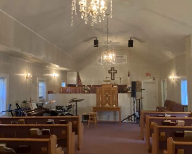 The interior of a church features wooden pews, a central pulpit with a cross, and musical instruments, including a piano and drums, under a chandelier.