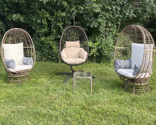 The image shows three woven garden chairs, including a hanging chair, arranged on a grassy area surrounded by trees.