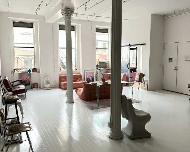 The image shows a spacious, modern interior with white floors, large windows, and a mix of seating options, including sofas and chairs, alongside decorative columns.