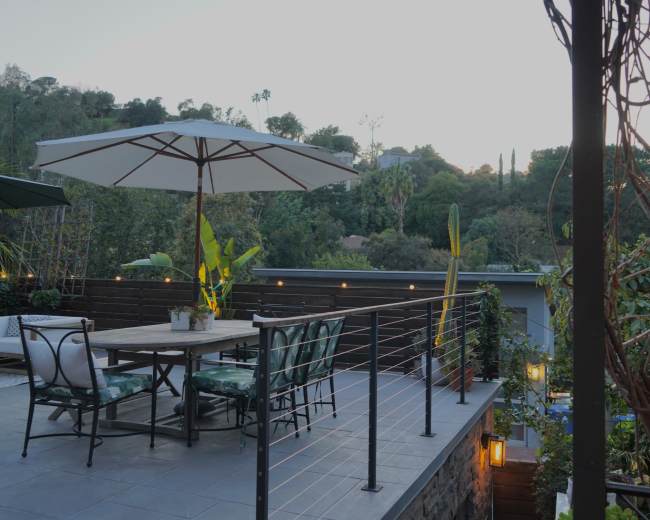 A patio with a dining table and chairs is surrounded by plants and features an umbrella, overlooking a wooded hillside at dusk.