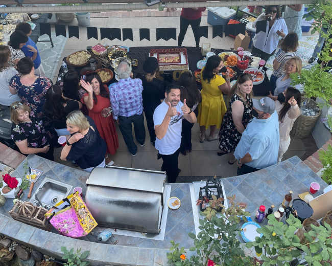 A crowd gathers around a buffet table at an outdoor gathering under a metal roof.