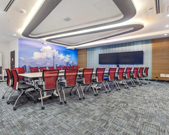 The image shows a modern conference room featuring a long table surrounded by red mesh chairs, with a large screen at one end and a cityscape mural on the wall.