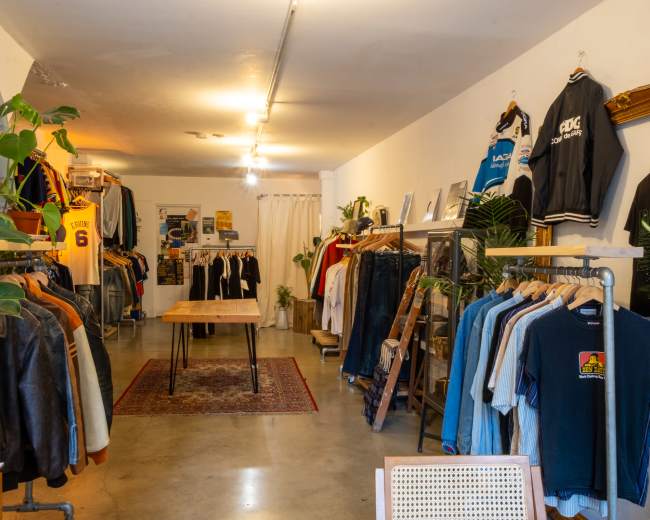 The image shows the interior of a clothing boutique featuring racks of vintage apparel and a wooden table in a well-lit space.