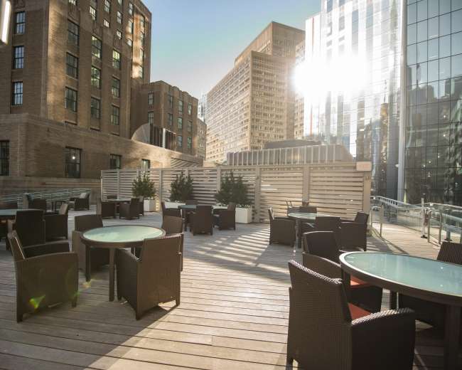 The image shows an outdoor terrace featuring several round tables surrounded by wicker chairs, with modern buildings in the background and sunlight reflecting off the glass surfaces.