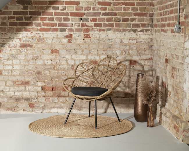 A woven rattan chair with a dark cushion sits on a round jute rug against a brick wall, accompanied by decorative vases and dried plants.