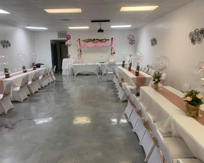 The image shows a decorated event space featuring several tables with white tablecloths and floral centerpieces arranged neatly along a polished concrete floor.