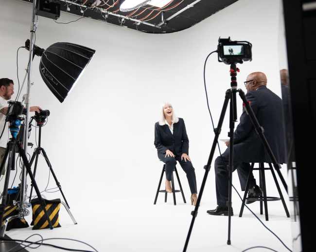 A woman in a black suit laughs while sitting on a stool during a film shoot in a studio with white walls and multiple cameras and lights set up.