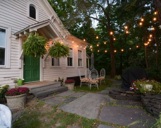 A cozy outdoor seating area with string lights illuminates the entrance of a house surrounded by greenery and well-maintained flower pots.