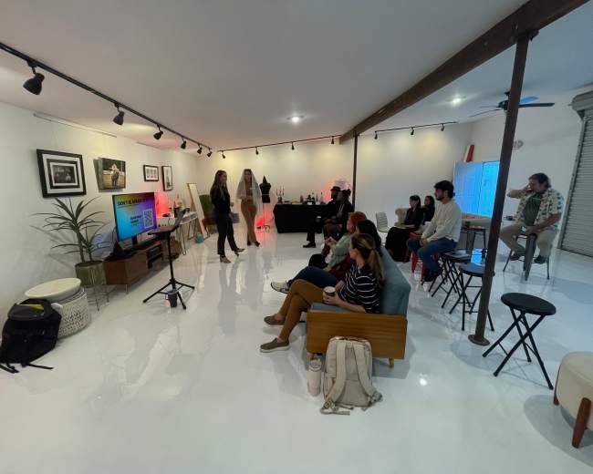 A group of people sits and stands in a brightly lit room, facing a large screen while engaging in an event.