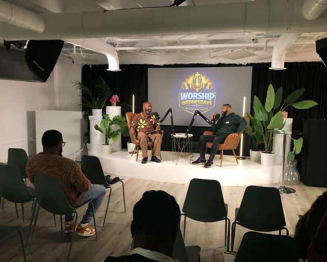 Two men are seated in stylish chairs on a stage with a decorative backdrop for an event labeled "Worship Wednesdays," while an audience sits in chairs facing them.