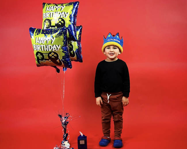 A child stands next to three colorful birthday balloons against a red background, wearing a crown and smiling.