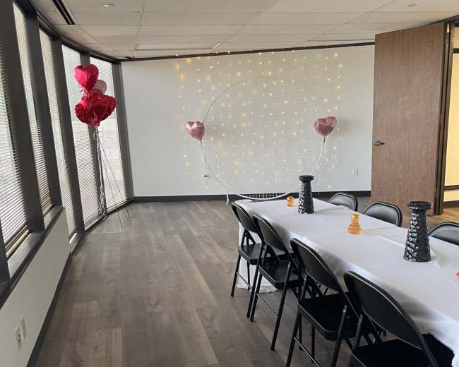 A long table with black chairs is set up in a brightly lit room, featuring a backdrop of twinkling fairy lights and heart-shaped balloons on either side.