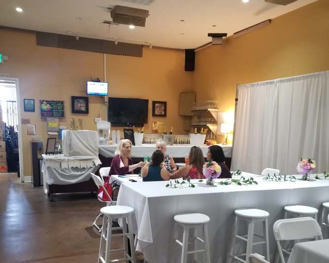 Four women are seated at a table in a brightly lit event space, surrounded by white tablecloths and floral centerpieces.