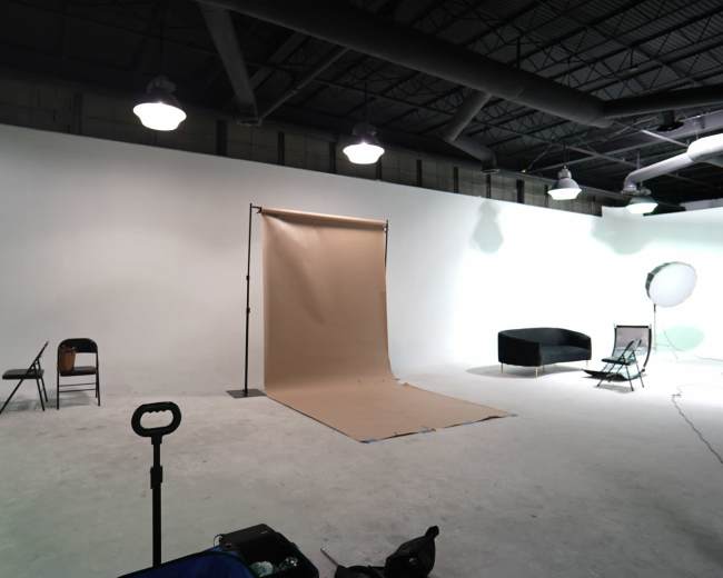 A photography studio with a beige backdrop set up on a wooden floor, surrounded by several empty chairs and a couch.