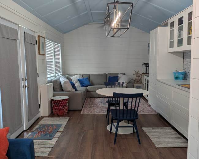 The image shows the interior of a small, modern cabin featuring a living area with a sectional sofa, a dining table with chairs, and a kitchen area with white cabinetry.