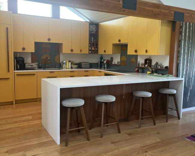 The image shows a modern kitchen featuring yellow cabinetry, a central white island, and wooden bar stools.