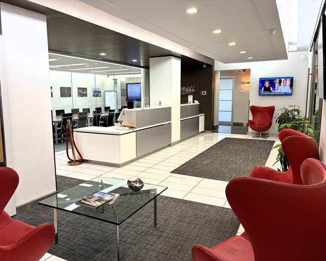 The image shows a modern office lobby featuring red chairs, a glass coffee table, and a reception area with a waiting area and meeting rooms visible in the background.