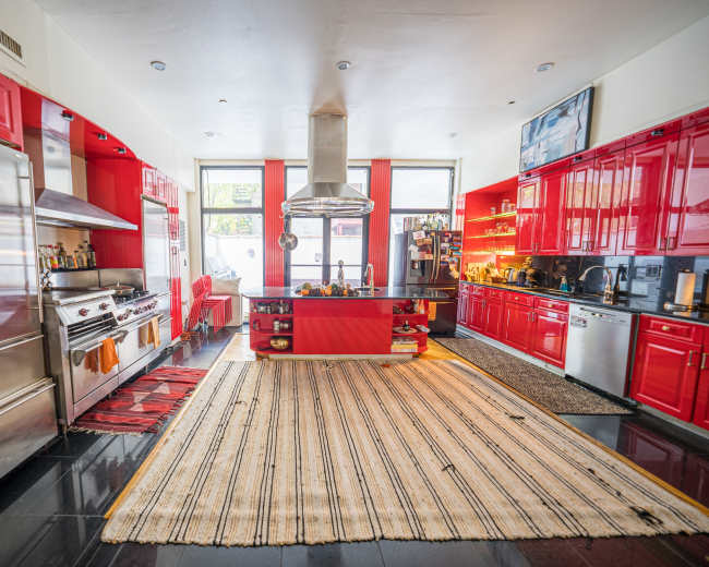 A spacious kitchen features red cabinetry, stainless steel appliances, and a central island with a polished surface, surrounded by patterned rugs.