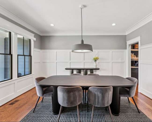 A modern dining room features a long black table surrounded by gray upholstered chairs, with a pendant light hanging above and a built-in cabinet visible in the background.