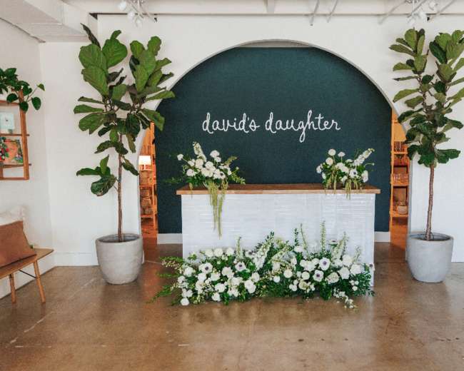 The image shows a reception area with a white desk against a dark green wall, adorned with the words "david's daughter," surrounded by large potted plants and white floral arrangements.