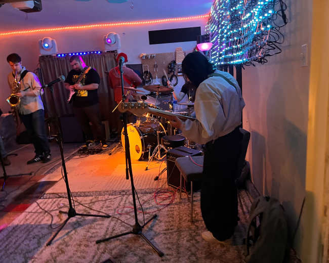 A band performs on a small stage with colorful lights and musical instruments in a cozy room.