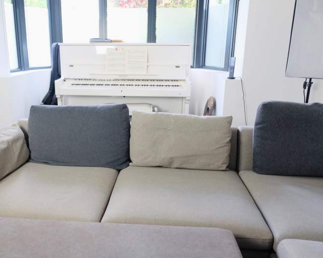 A light-colored sectional sofa sits in a room with large windows, adjacent to a white piano.