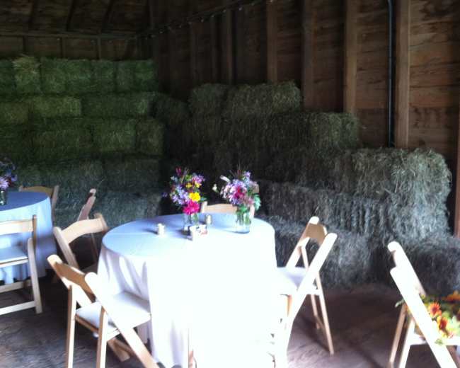 The scene features a rustic interior with stacked hay bales against the wall and two round tables adorned with white tablecloths and floral arrangements.