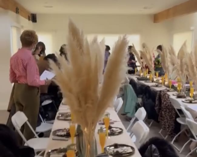 A long table is set with decorative plates and glasses, flanked by tall pampas grass arrangements in a brightly lit indoor space filled with people.