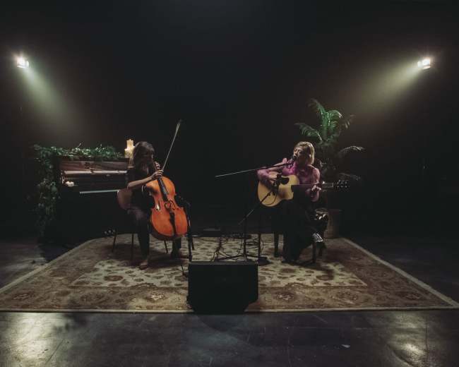 Two musicians are performing together, one playing a cello and the other an acoustic guitar, in a dimly lit room with candles and plants.