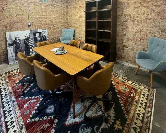 A meeting room features a large wooden table surrounded by brown chairs, with a patterned rug on the floor and a bookshelf against a brick wall.
