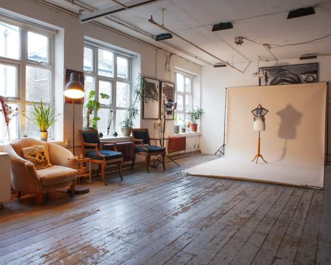 The image shows a spacious studio with large windows, featuring a seating area, plants, and a photography backdrop set up for a photoshoot.