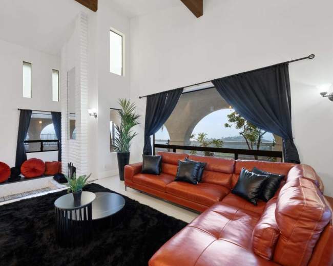 The image features a modern living room with a red leather sofa, dark curtains, a black coffee table, and a view of greenery through large windows.