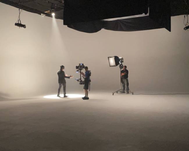 A film crew is set up in a studio, with one person speaking under a spotlight while a camera operator films them.