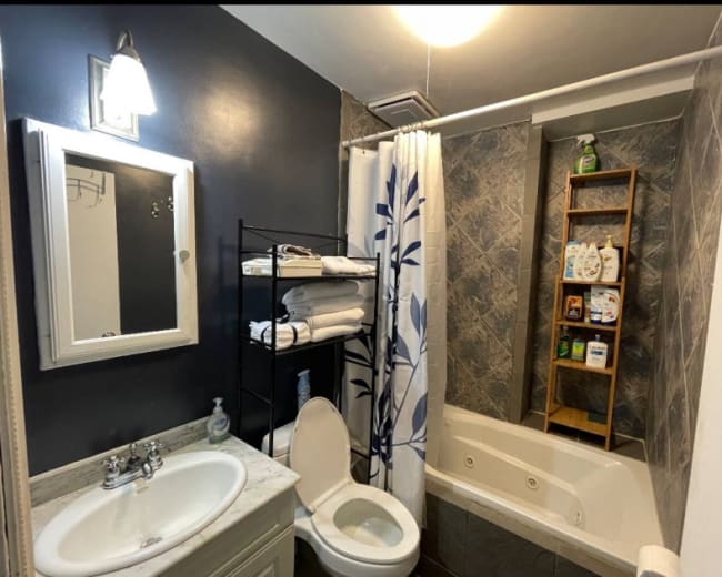 A compact bathroom featuring a tub, a toilet, a mirrored cabinet, and a shelving unit with neatly arranged toiletries.