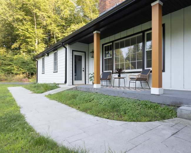 The image shows a house with a porch, featuring two chairs and a table, and a path made of stone leading up to the entrance.
