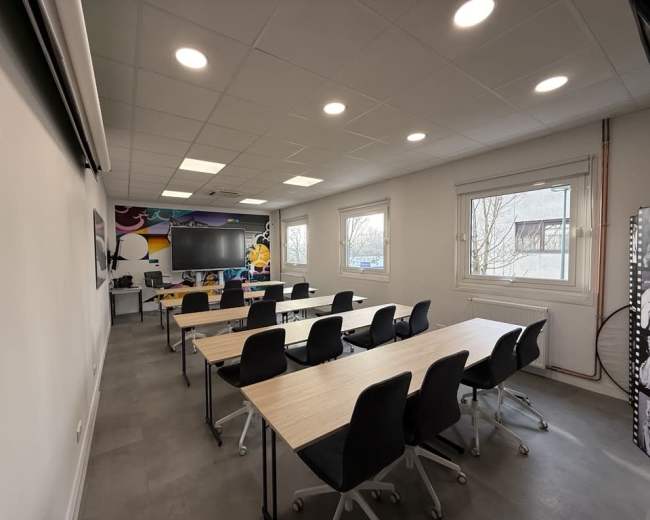 The image shows a modern classroom with multiple tables arranged in rows, black chairs, and large windows allowing natural light, along with a wall featuring colorful murals.