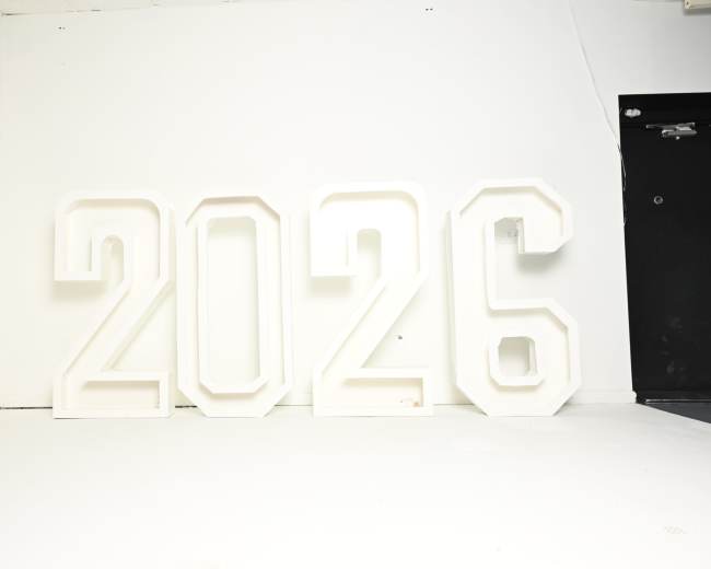 The image shows large, white illuminated numbers "2026" arranged against a plain white wall.