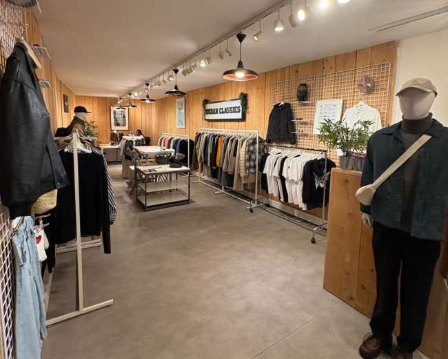 The image shows a clothing store interior with racks of apparel on either side, wooden walls, and a central display table under overhead lighting.