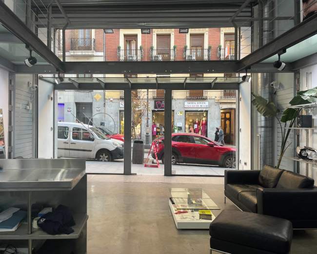 The image shows a modern interior space with a view of a street through large glass doors, featuring a black sofa, a coffee table, and parked vehicles outside.