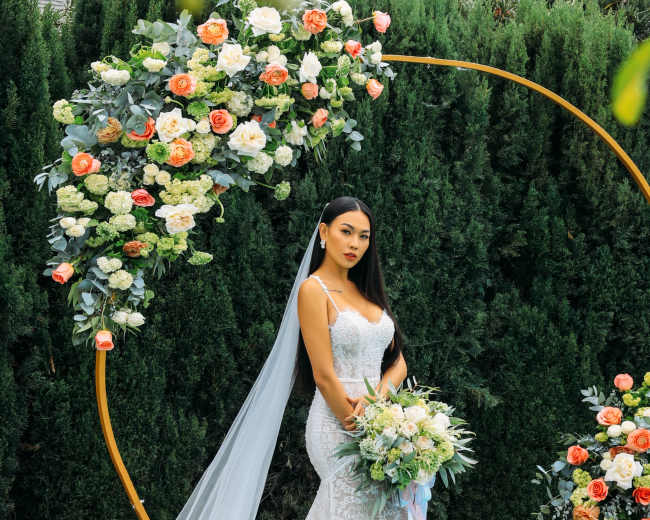 A bride in a fitted lace gown poses with a bouquet in front of a floral arch adorned with roses and greenery.