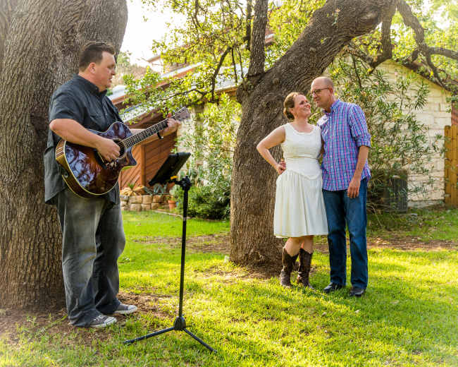 A man plays guitar while a couple poses affectionately together under two large trees in a grassy area.