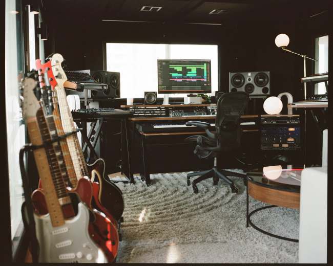 The image shows a modern music production studio featuring several guitars on stands, a central workstation with a computer and audio equipment, and large speakers in a sleek, well-lit space.