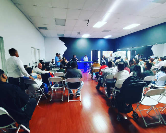 A large gathering of people sits in rows of chairs facing a panel of speakers at a community event.