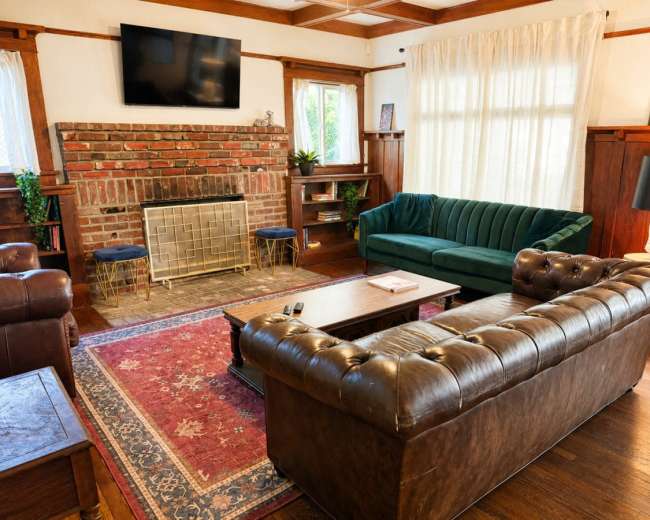 The image shows a cozy living room featuring a brick fireplace, a green velvet sofa, and a brown leather chesterfield couch, with wooden accents and a patterned rug.