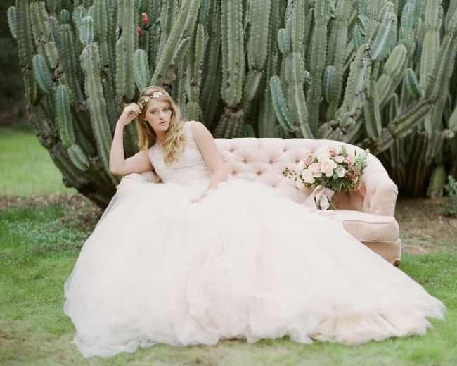 A bride in a flowing gown sits on a light pink couch beside a large cactus in a green outdoor setting.