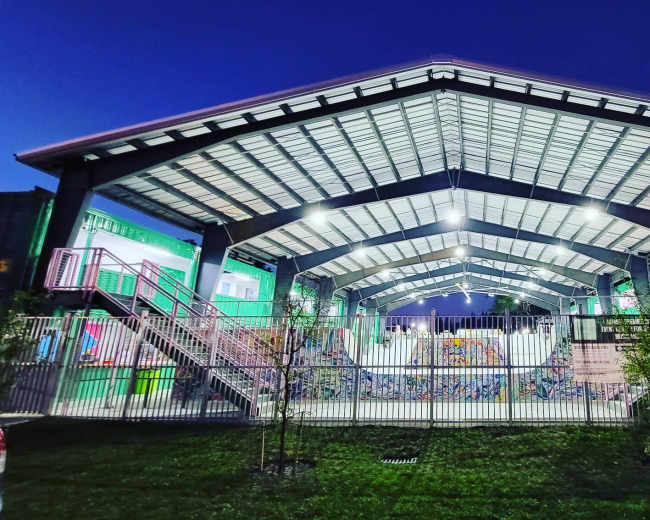 A large, open-air structure with a peaked roof houses a recreational area, illuminated under evening lights, with colorful murals on the walls.